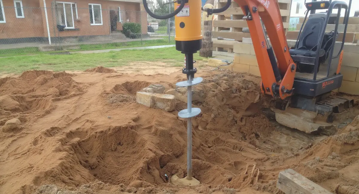 Torque motor screwing a helical pier shaft with round helix plates into brown clay soil at a residential foundation, with multiple installed piers visible in background